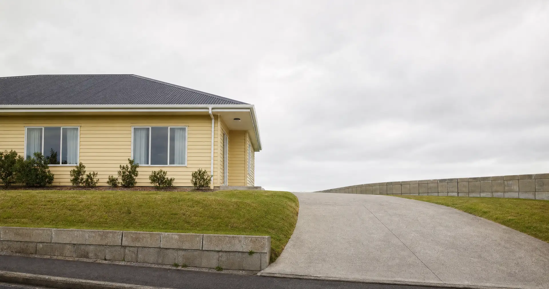 A single-storey pale yellow weatherboard house with a dark iron roof. A concrete driveway and a mown berm run in front.